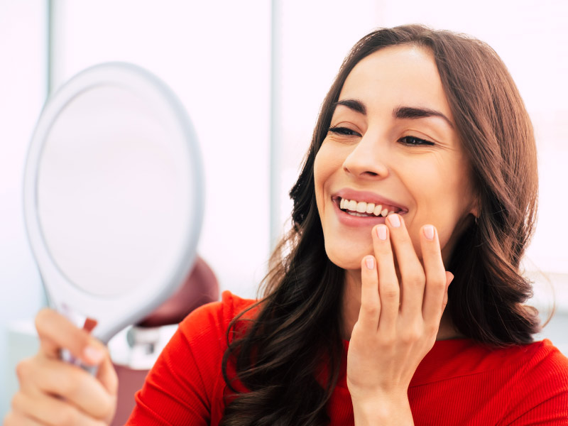 Brunette woman looks in handheld mirror to admire her tooth replacements after missing teeth