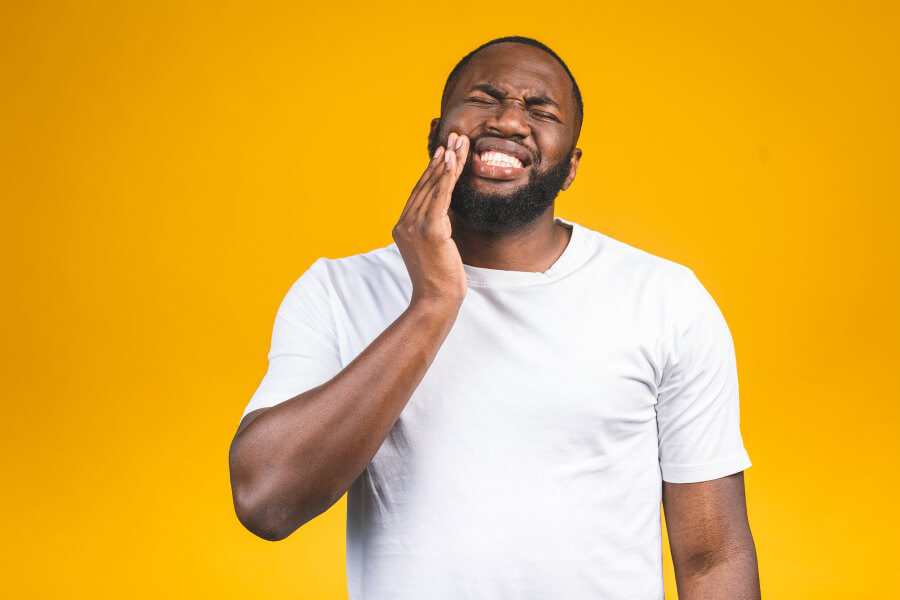 Man in white shirt cringes in pain and touches his cheek due to a toothache dental emergency