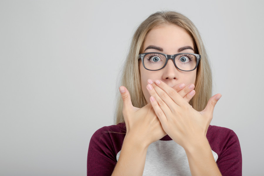 young woman wearing glasses covers her mouth with both hands to hide gum disease