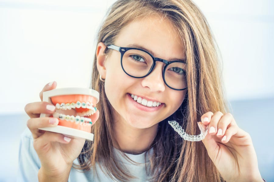 Smiling teen holding a mouth model wearing traditional braces in one hand and an Invisalign clear aligner in the other.