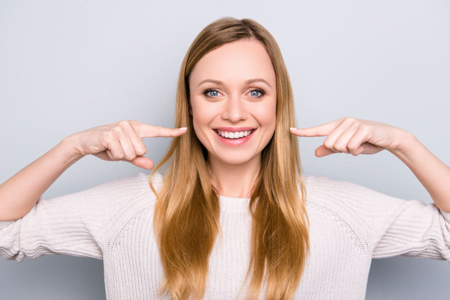 A smiling woman with long blond hair pointing at her white teeth.