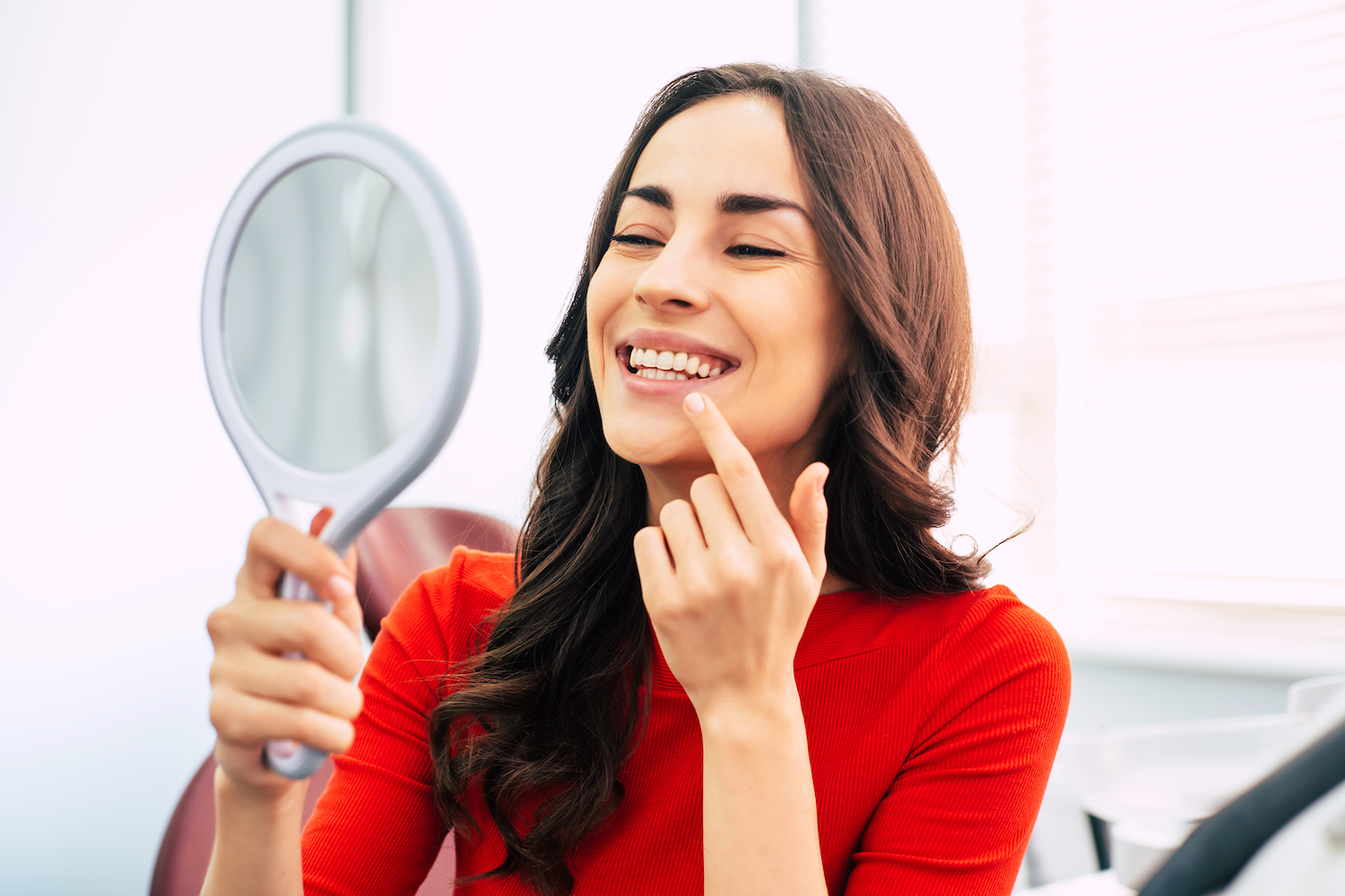 brunette woman in a red blouse smiles at her teeth in a handheld mirror after a smile makeover