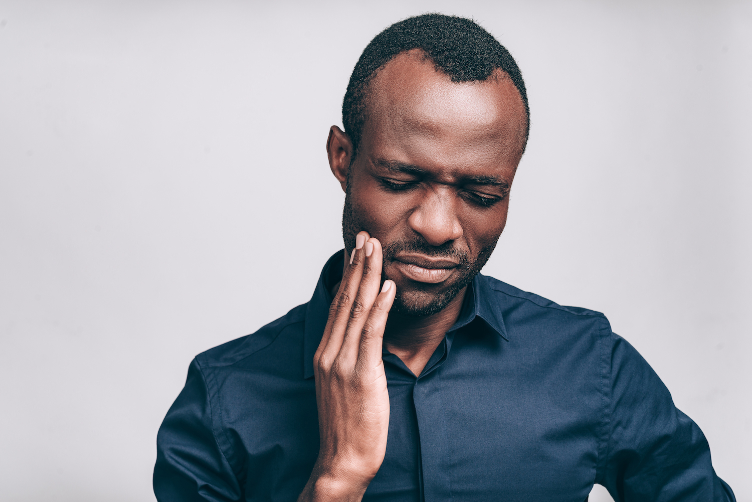 Black man in a blue dress shirt cringes in pain and touches his cheek wondering if he'll need root canal therapy