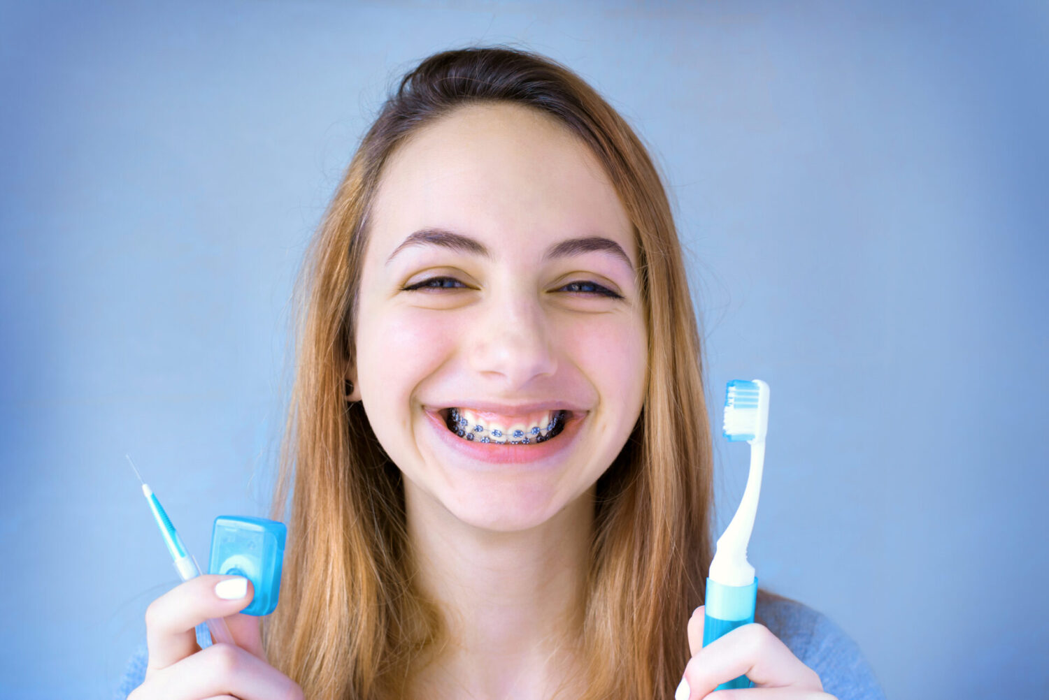 Blonde girl smiles with braces while holding a toothbrush, a floss container, and an interdental brush